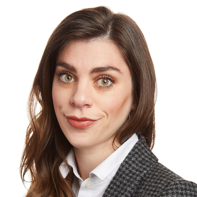 Close-up headshot of a woman with long brown hair, hazel-green eyes, subtle smile, wearing a white collared shirt and gray blazer.