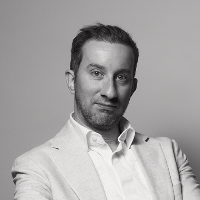 Black-and-white headshot: short hair, light stubble, white shirt, light blazer, arms crossed, neutral expression on plain gray background.