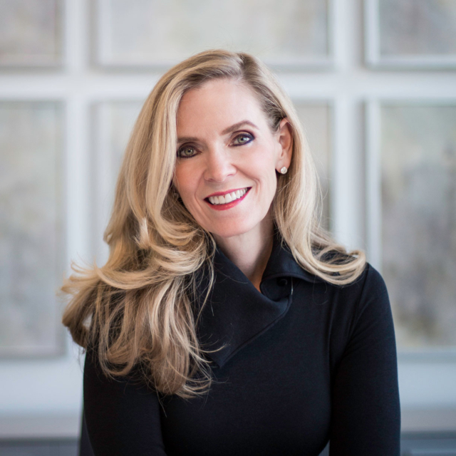 A woman with long blonde hair and a warm smile wears a black top, posing against a softly blurred, light-colored background.