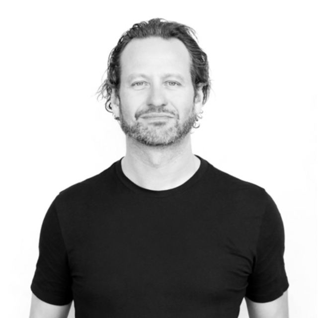 A man with wavy hair and a beard stands confidently, wearing a black t-shirt against a plain white background. He appears relaxed and approachable.