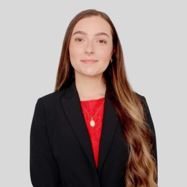 A young woman with long, wavy hair wears a black blazer over a red top, smiling confidently against a light gray background.