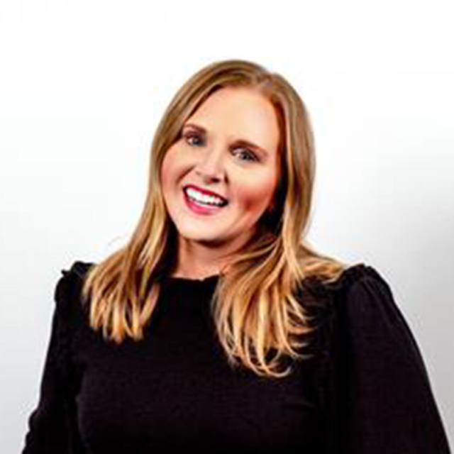 Smiling fair-skinned woman with shoulder-length blonde hair wearing a black long-sleeve top, head tilted slightly against a pale background.
