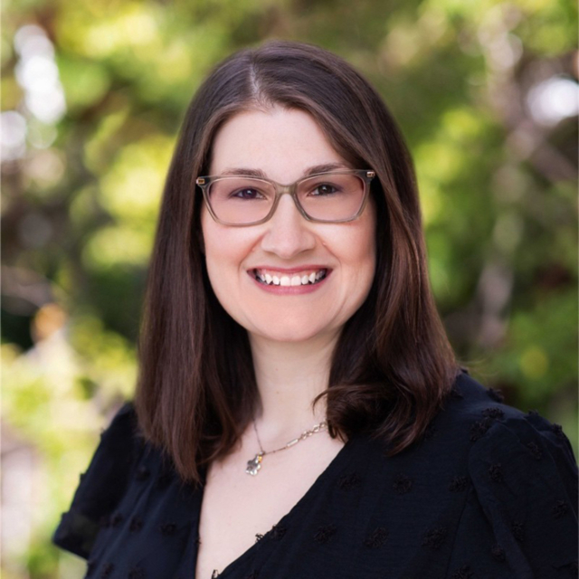 A smiling woman with long dark hair and glasses poses outdoors, wearing a black top and a necklace, against a blurred green background.