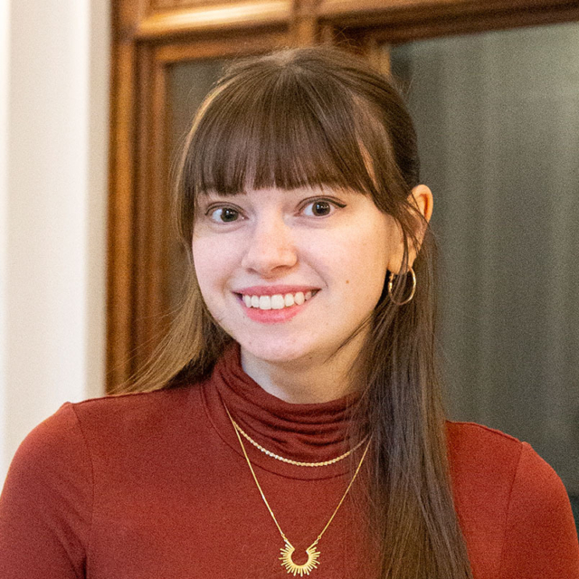 Smiling woman with long brown hair and bangs in a maroon turtleneck, wearing layered gold necklaces with sunburst pendant and hoop earrings.