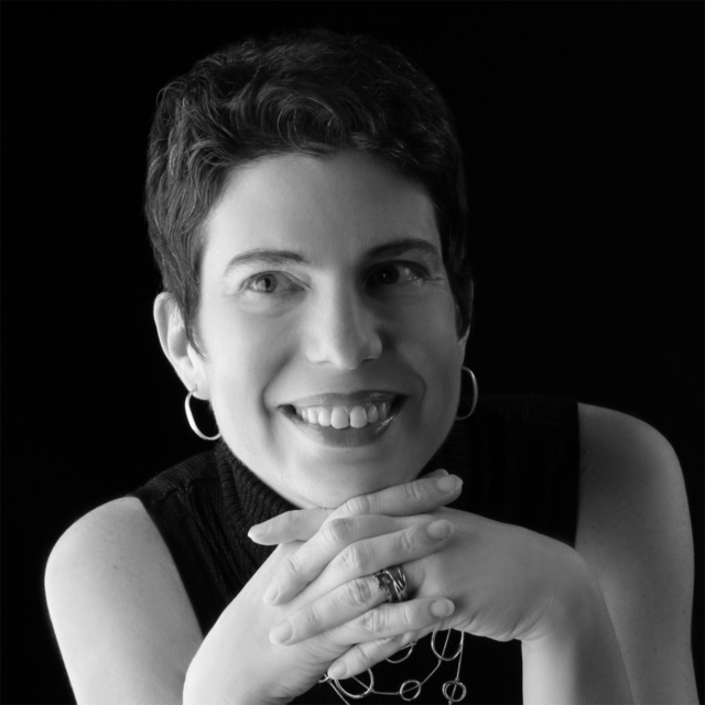 A smiling woman with short hair and hoop earrings rests her hands together, showcasing a friendly demeanor against a dark background.