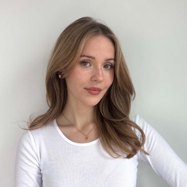 A young woman with long, wavy brown hair and soft makeup poses against a neutral background, wearing a white long-sleeve shirt.