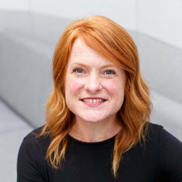 A smiling woman with wavy red hair and blue eyes wears a black top, sitting against a light gray background. The expression is warm and inviting.