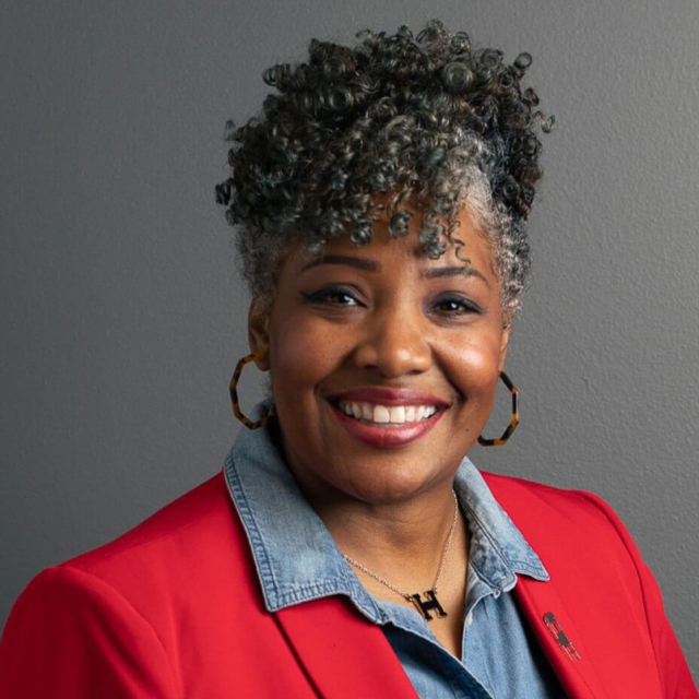 Smiling person with short gray-black curly hair, tortoiseshell hoop earrings, denim shirt under a red blazer and an H pendant necklace.