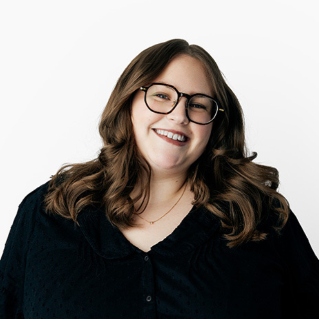Smiling woman with wavy shoulder-length brown hair and black-rimmed glasses wearing a dark blouse and gold necklace on a white background.