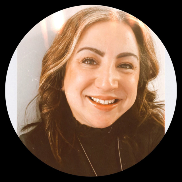 A smiling woman with wavy brown hair and subtle highlights poses against a light background, wearing a black top and necklace.