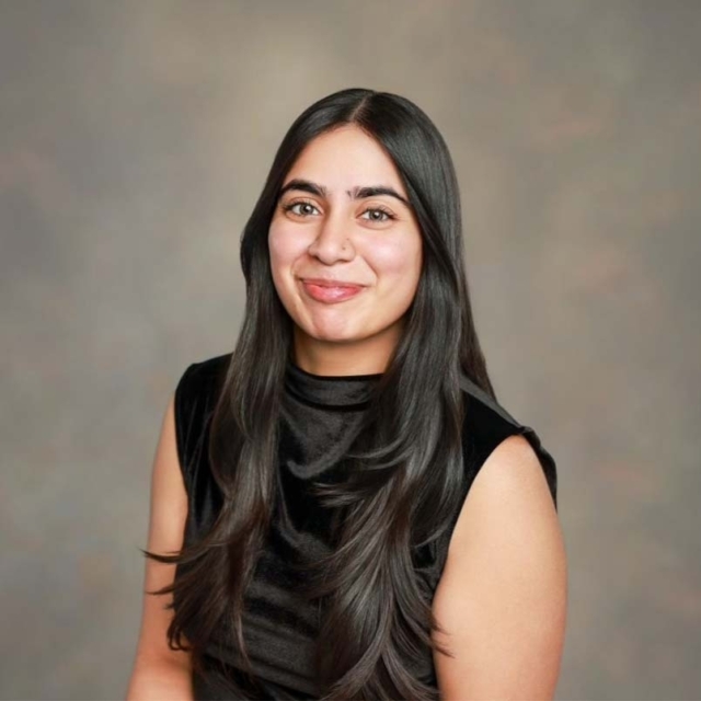A young woman with long, black hair smiles warmly while wearing a black sleeveless top against a softly blurred background.