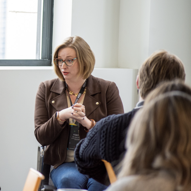 A woman with short blonde hair and glasses speaks animatedly during a workshop, engaging an audience in a bright, modern setting.