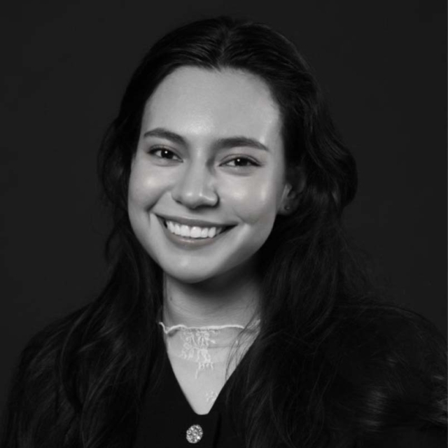A young woman with long, dark hair smiles warmly, wearing a black outfit with a delicate white collar against a dark background.