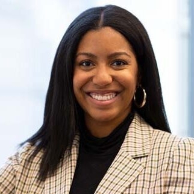 Smiling woman with long dark hair, wearing gold hoop earrings, black turtleneck and plaid blazer against a blurred indoor background.