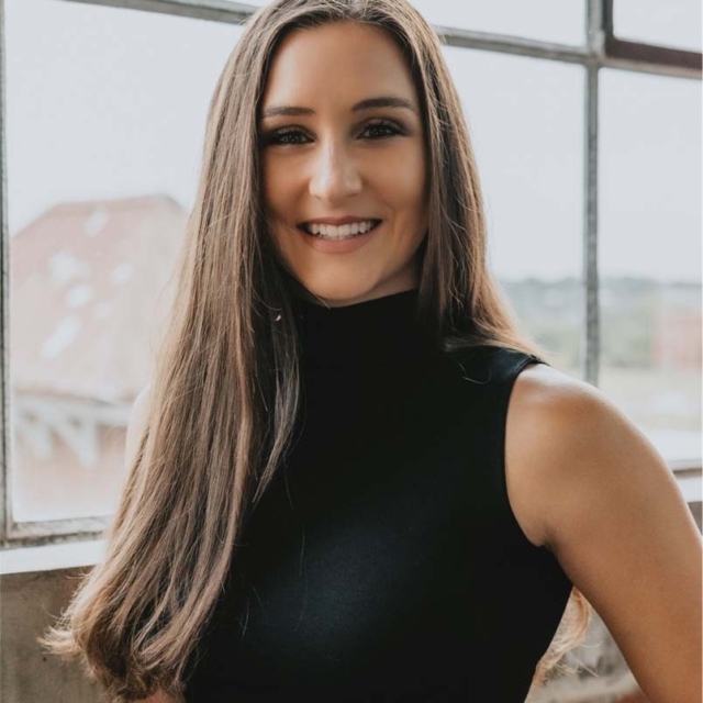 A young woman with long, straight hair smiles warmly, wearing a sleeveless black top against an industrial backdrop with large windows.