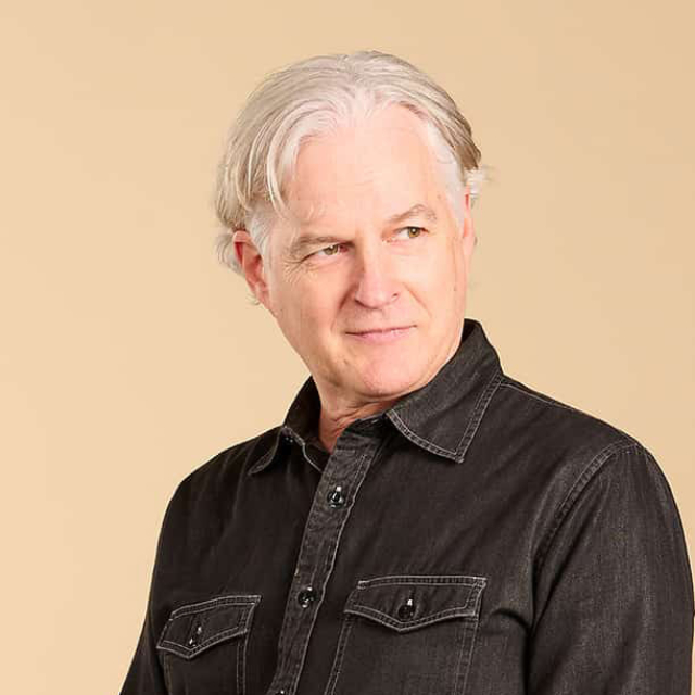A man with gray hair and a slight smile stands confidently, wearing a dark button-up shirt against a neutral background.