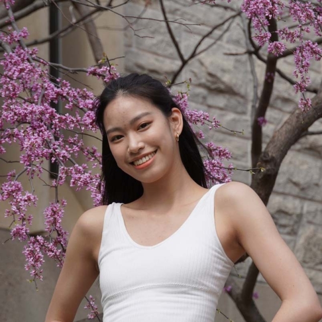 A young woman stands smiling in front of flowering pink branches, wearing a white tank top against a textured stone background.