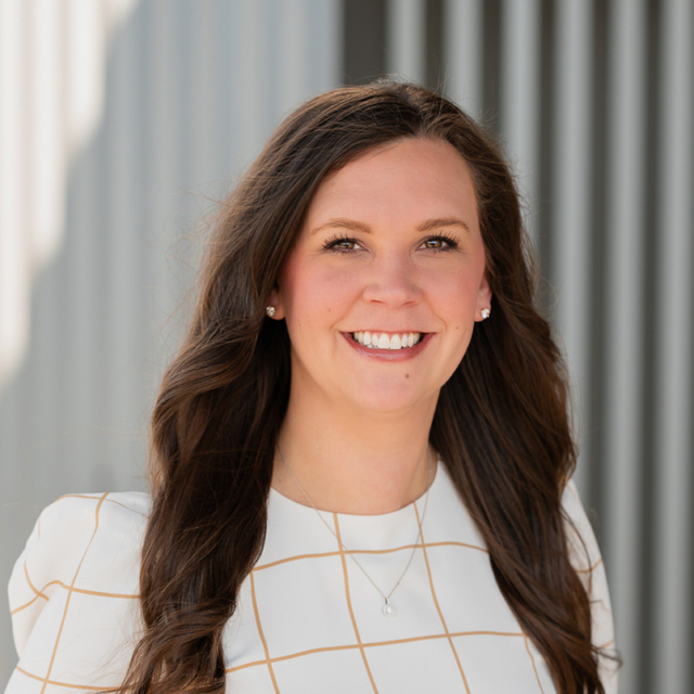 A woman with long, wavy brown hair smiles warmly while wearing a white blouse with a grid pattern, set against a textured background.