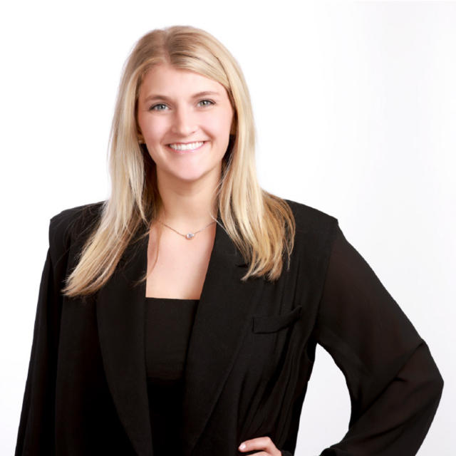 A smiling young woman with long blonde hair wears a black blazer and a black top, posing confidently against a white background.