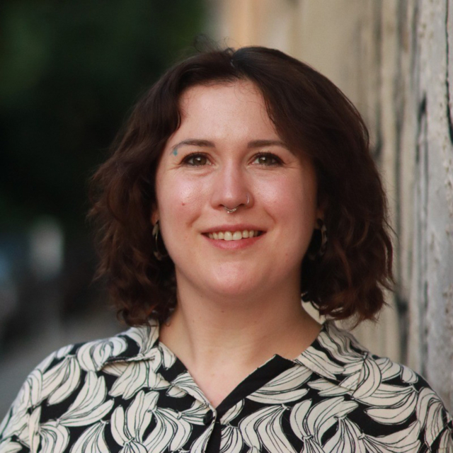 A person with curly hair and a septum piercing smiles warmly, wearing a leaf-patterned shirt against a blurred outdoor backdrop.