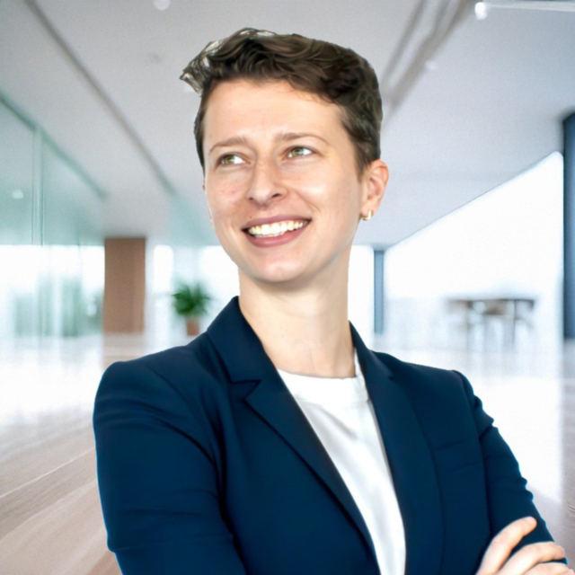 Smiling professional with short hair wearing a navy blazer and white shirt, standing confidently in a modern office space.