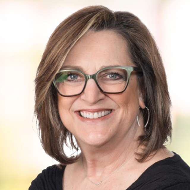 Smiling woman with shoulder-length brown hair, green glasses, and a black top, set against a blurred light background.