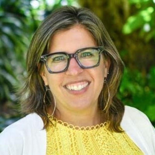 Smiling woman with shoulder-length brown hair and black-rimmed glasses wearing yellow lace top and white cardigan in leafy green backdrop.