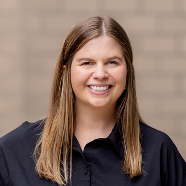 Smiling woman with straight shoulder-length light brown hair center-parted, in a black collared shirt against a brick background.