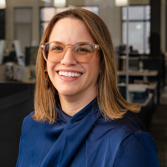 A smiling woman with shoulder-length brown hair wears glasses and a navy blouse, set against a modern office background.