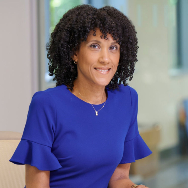 A woman with curly hair smiles while wearing a blue dress with ruffled sleeves, seated in a bright, modern office space.