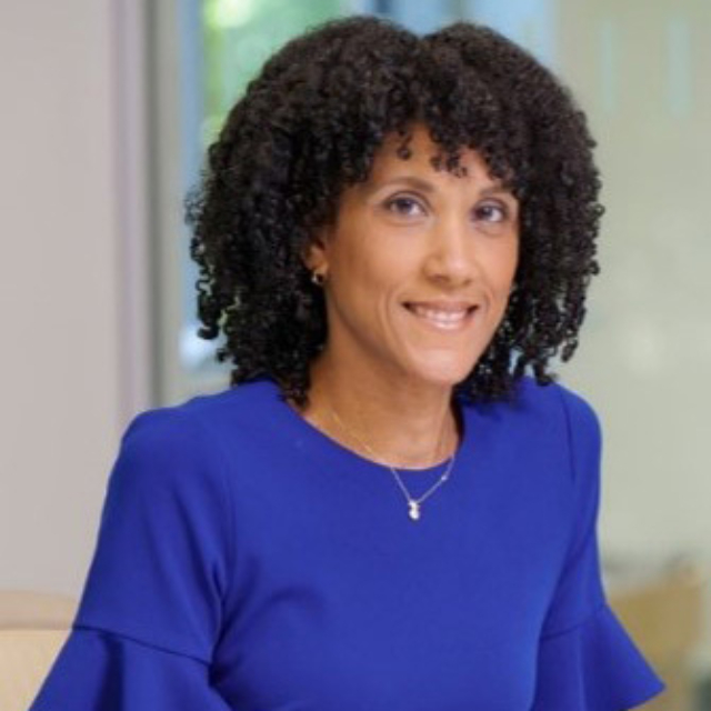A woman with curly hair wears a blue top with ruffled sleeves, smiling warmly while seated in a professional setting.