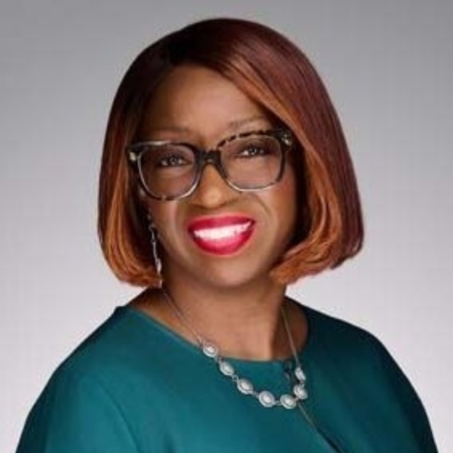 Smiling woman with short auburn bob, wearing black-rimmed glasses, teal dress and silver necklace against soft gray backdrop.