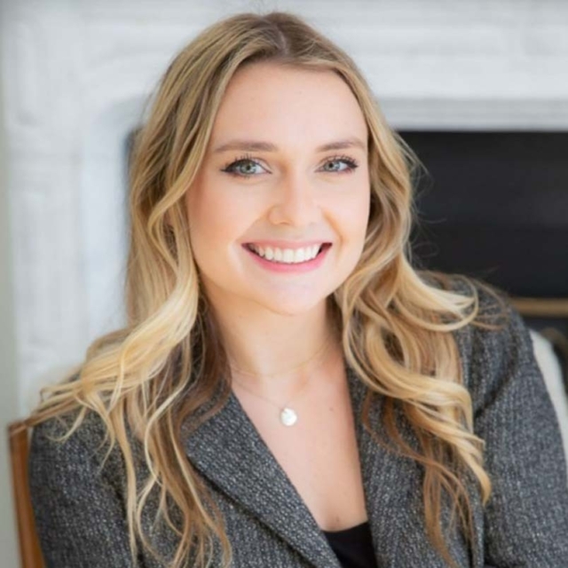 A young woman smiles warmly, showcasing long, wavy blonde hair and wearing a stylish gray blazer, against a softly blurred background.