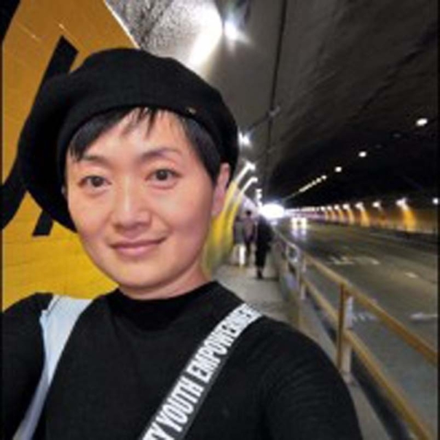 A woman with short hair and a beret stands in a brightly lit tunnel, smiling at the camera with traffic in the background.
