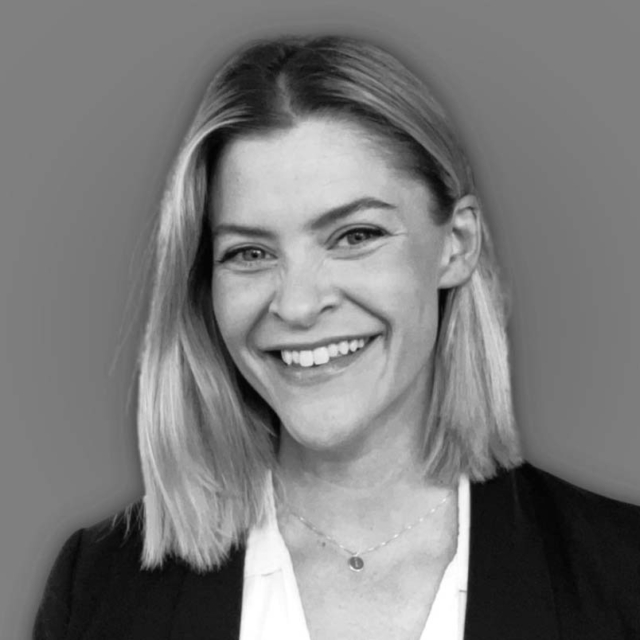 A smiling woman with shoulder-length blonde hair and a black blazer poses against a neutral gray background. Her expression is warm and inviting.