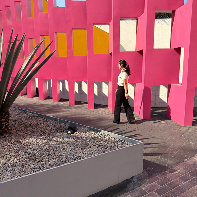 A woman in black pants walks beside a vibrant pink wall with cut-out squares, accented by a small agave plant and pebbled ground.