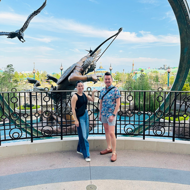 Two people stand beside a large bronze statue in an amusement park, with greenery and attractions visible in the background.