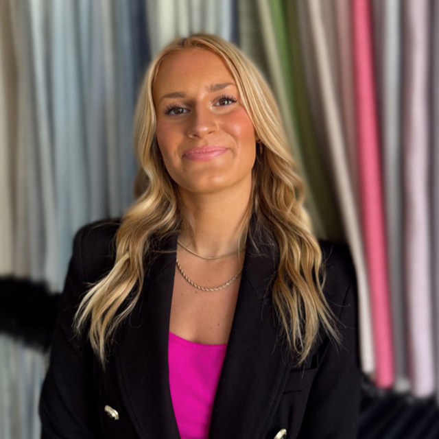 A young woman with long blonde hair smiles confidently, wearing a black blazer over a pink top, with colorful fabric rolls in the background.