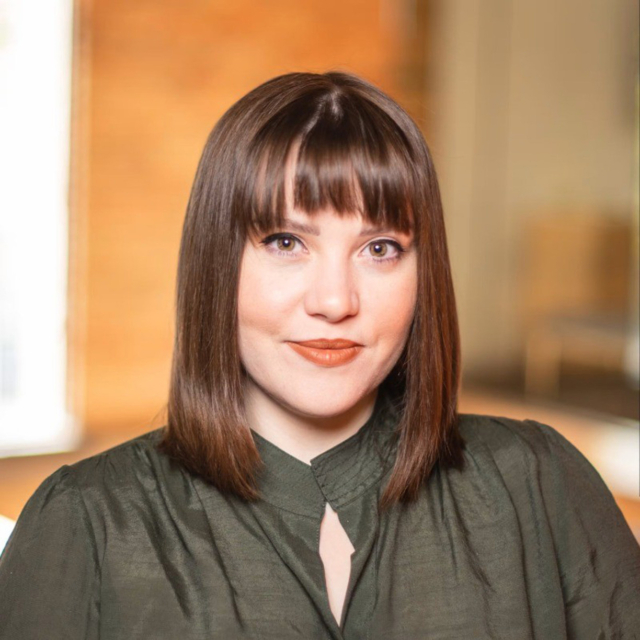 A woman with shoulder-length brown hair and bangs wears a green blouse, posing confidently with a soft background of warm tones.