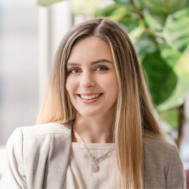 A woman with long, straight hair smiles warmly, wearing a light-colored blazer and necklace, with green plants in the background.