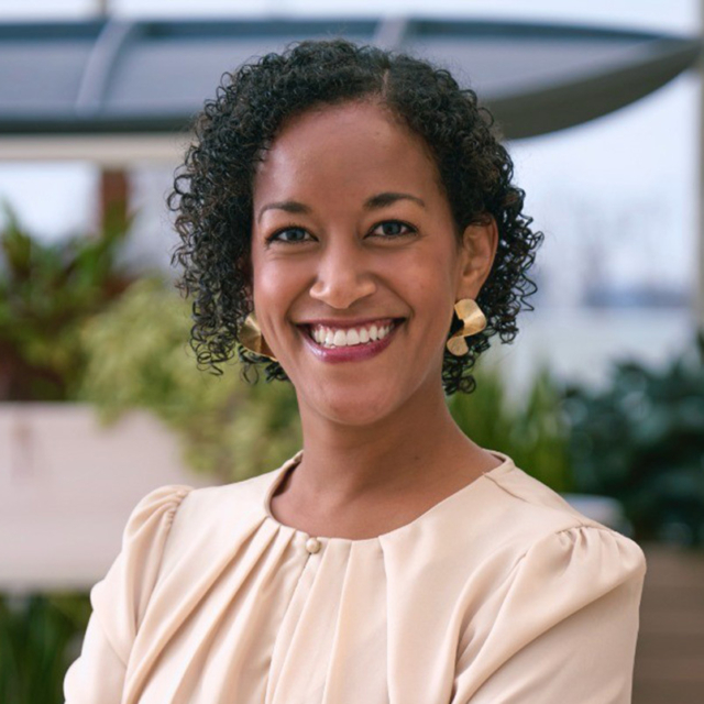 Smiling woman with short curly hair, gold geometric earrings and a cream blouse, standing in bright office space with blurred plants.