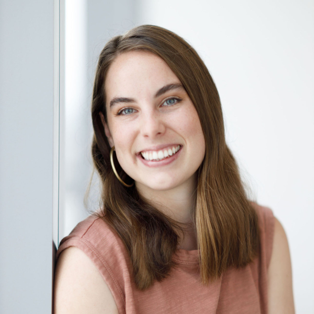 A young woman with long brown hair smiles brightly, wearing a rust-colored top and hoop earrings, against a soft, light backdrop.