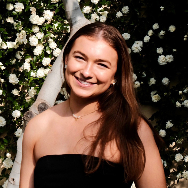 Young woman with long brown hair and pearl earrings smiling in a strapless black top, standing before a trunk and white camellia blossoms.