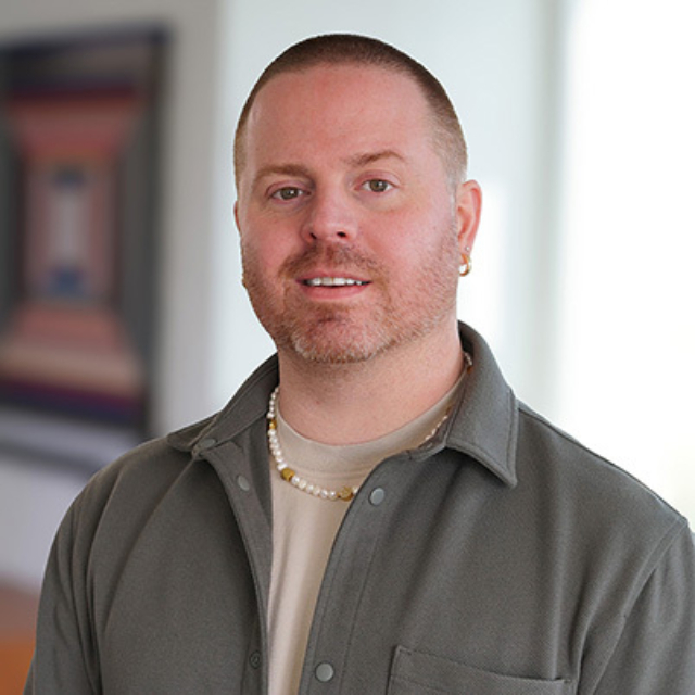 A man with short hair and a beard stands in a bright indoor space, wearing a light shirt and an oversized jacket, smiling warmly.
