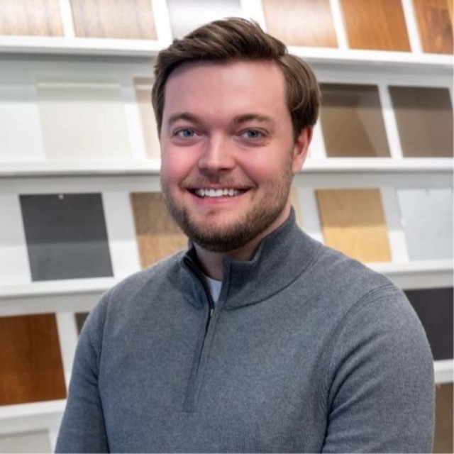 A smiling man with short brown hair and a beard wears a gray sweater, standing in front of a wall displaying various paint and wood samples.