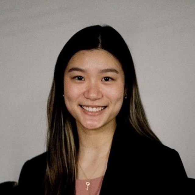 A young woman smiles warmly, wearing a black blazer over a pink top, with long hair and a key necklace against a neutral background.