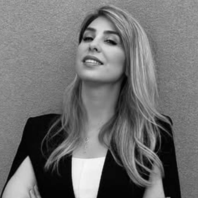 A confident woman with long, wavy hair is posing with her arms crossed, wearing a dark blazer against a neutral backdrop.