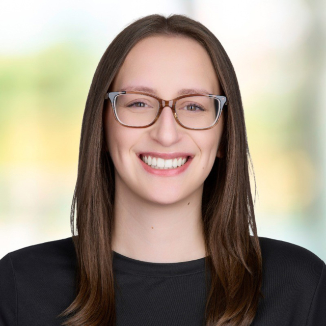 A smiling woman with long brown hair and glasses, wearing a black shirt, stands against a softly blurred background.