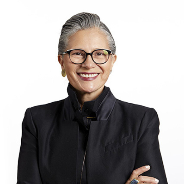 Smiling older woman with slicked-back gray hair and black glasses, wearing gold earrings and a high-collared black jacket, arms crossed.