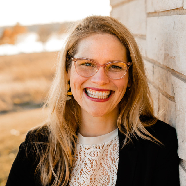 A smiling woman with long, blonde hair and glasses leans against a stone wall, showcasing a warm, inviting expression.